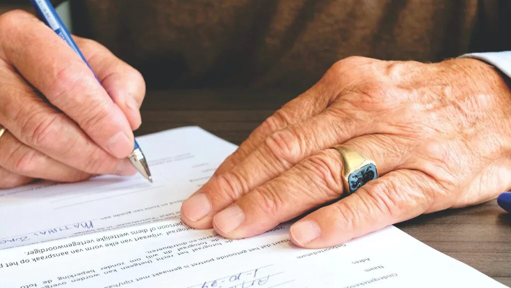 A close-up image of a person’s hands writing on a piece of paper with a pen. The left hand is holding the paper steady while the right hand writes. There is text and some mathematical expressions on the paper, but it is not fully visible to transcribe or identify its content. The person is wearing a ring on the left hand’s ring finger.