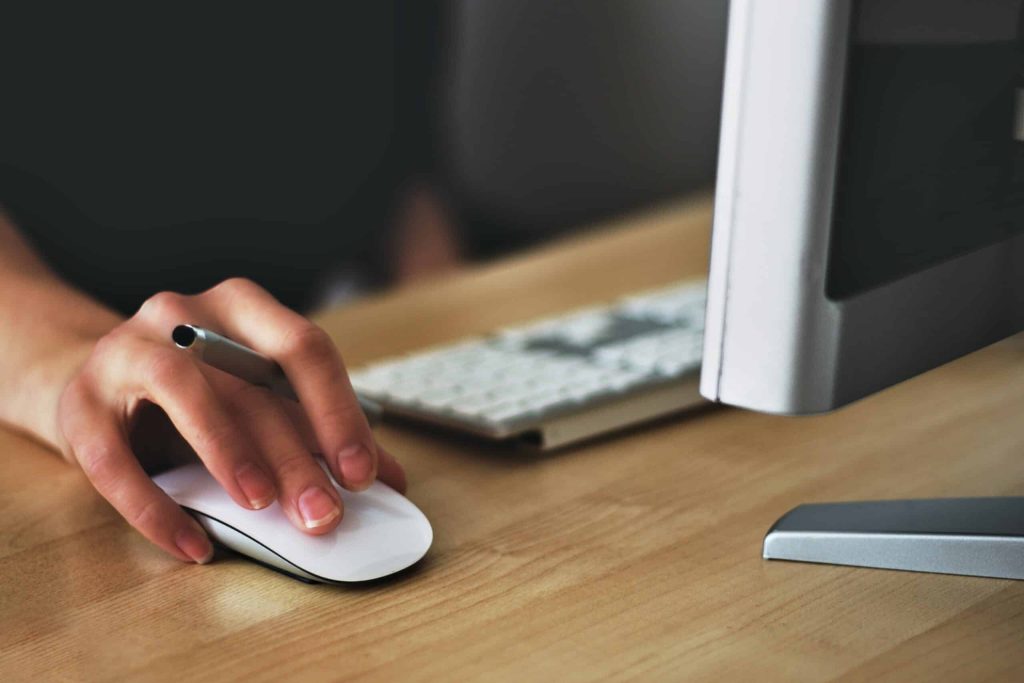 Close-up of a hand using a white computer mouse next to a keyboard and part of a monitor on a wooden desk. #Geraci Client Corner