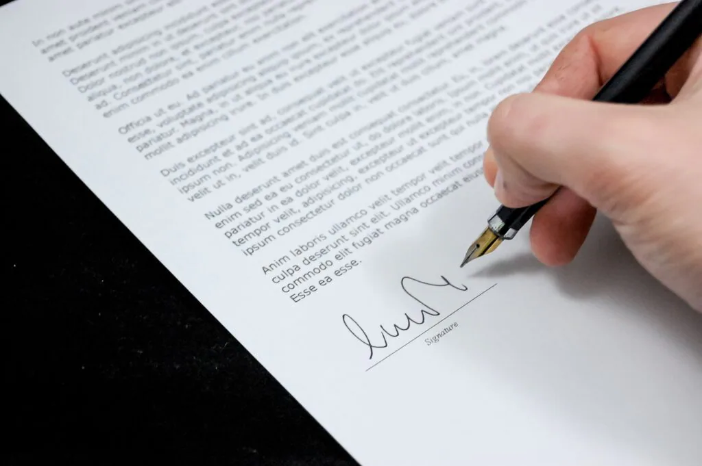 A close-up image of a person’s hand holding a black pen and signing a document. The document contains several paragraphs of text with the signature at the bottom, indicating the finalization of an agreement or contract.