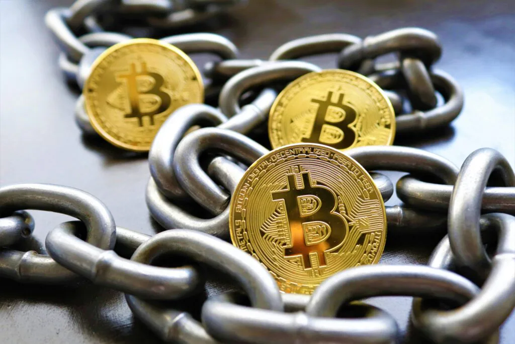 A close-up image of two shiny gold-colored Bitcoin tokens linked by a heavy metal chain. The foremost Bitcoin token is in sharp focus, showing the ‘B’ symbol with two vertical lines through it, indicative of the digital cryptocurrency. The background features a blurred second Bitcoin and additional loops of the chain, all resting on a dark surface that reflects some light. #Blockchain in Banking