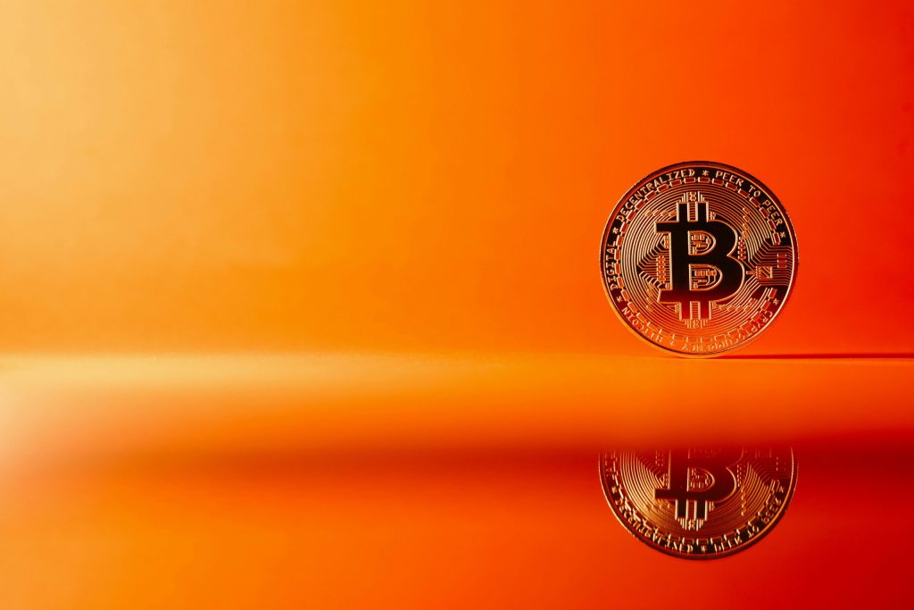 Blockchain in Banking: A single Bitcoin coin stands upright on a reflective surface against an orange background, symbolizing the digital currency’s role in the future of banking.