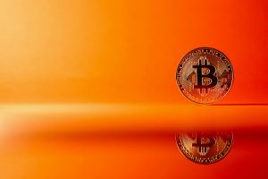 Blockchain in Banking: A single Bitcoin coin stands upright on a reflective surface against an orange background, symbolizing the digital currency’s role in the future of banking.
