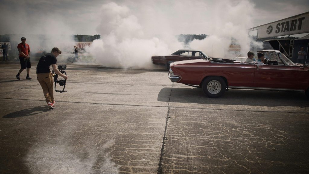 A red vintage car performing a burnout at a start line with white smoke billowing from the rear tires, as a person films the action.