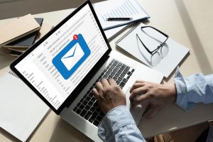 Person typing on a laptop with an email application open on the screen, surrounded by various office items including papers, a notebook, and eyeglasses. The man is working with Cliqly Email Marketing.