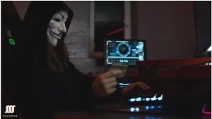 Where can I hire a hacker: A person wearing a white Guy Fawkes mask and a hooded cloak works at a desk with a glowing computer screen displaying technical data, holding a card, in a dimly lit room.