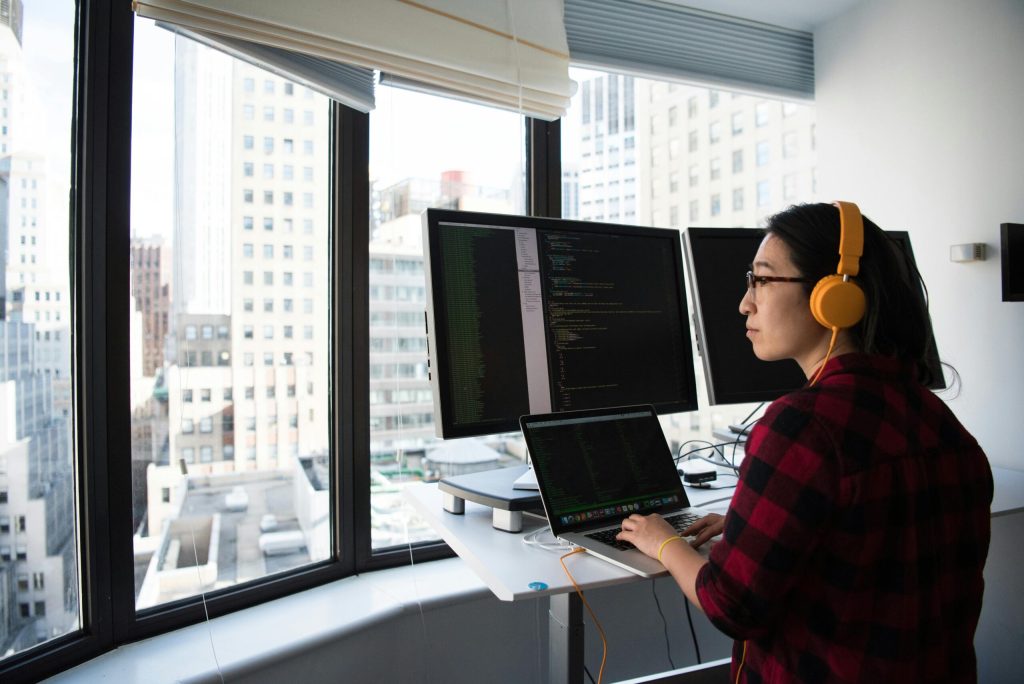 A person with obscured face working on multiple computer screens with code visible, in an office setting with a cityscape visible through the window. #ComputerVisionSyndrome