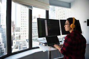 A person with obscured face working on multiple computer screens with code visible, in an office setting with a cityscape visible through the window. #ComputerVisionSyndrome