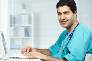 A male Medical doctor wearing teal scrubs and a stethoscope around his neck is sitting at a desk using a laptop. He is smiling and looking towards the camera.