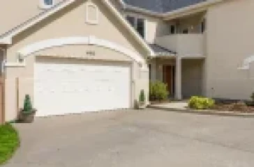 A modern two-story house with a beige exterior, white garage door, and large windows, featuring a spacious driveway and well-maintained landscaping with shrubs and a wooden fence.