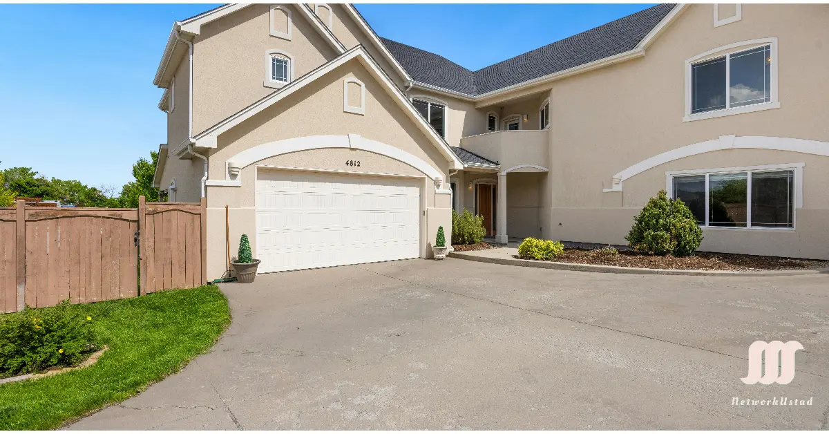 A modern two-story house with a beige exterior, white garage door, and large windows, featuring a spacious driveway and well-maintained landscaping with shrubs and a wooden fence.