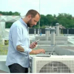 A man in a light blue shirt using a laptop and smartphone near an air conditioning unit on a rooftop, surrounded by industrial equipment and greenery.