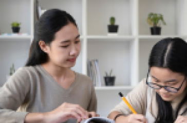 Two Asian women studying together at a table with books, notebooks, and a tablet in a modern room with white bookshelves and small potted plants in the background.