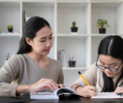 Two Asian women studying together at a table with books, notebooks, and a tablet in a modern room with white bookshelves and small potted plants in the background.