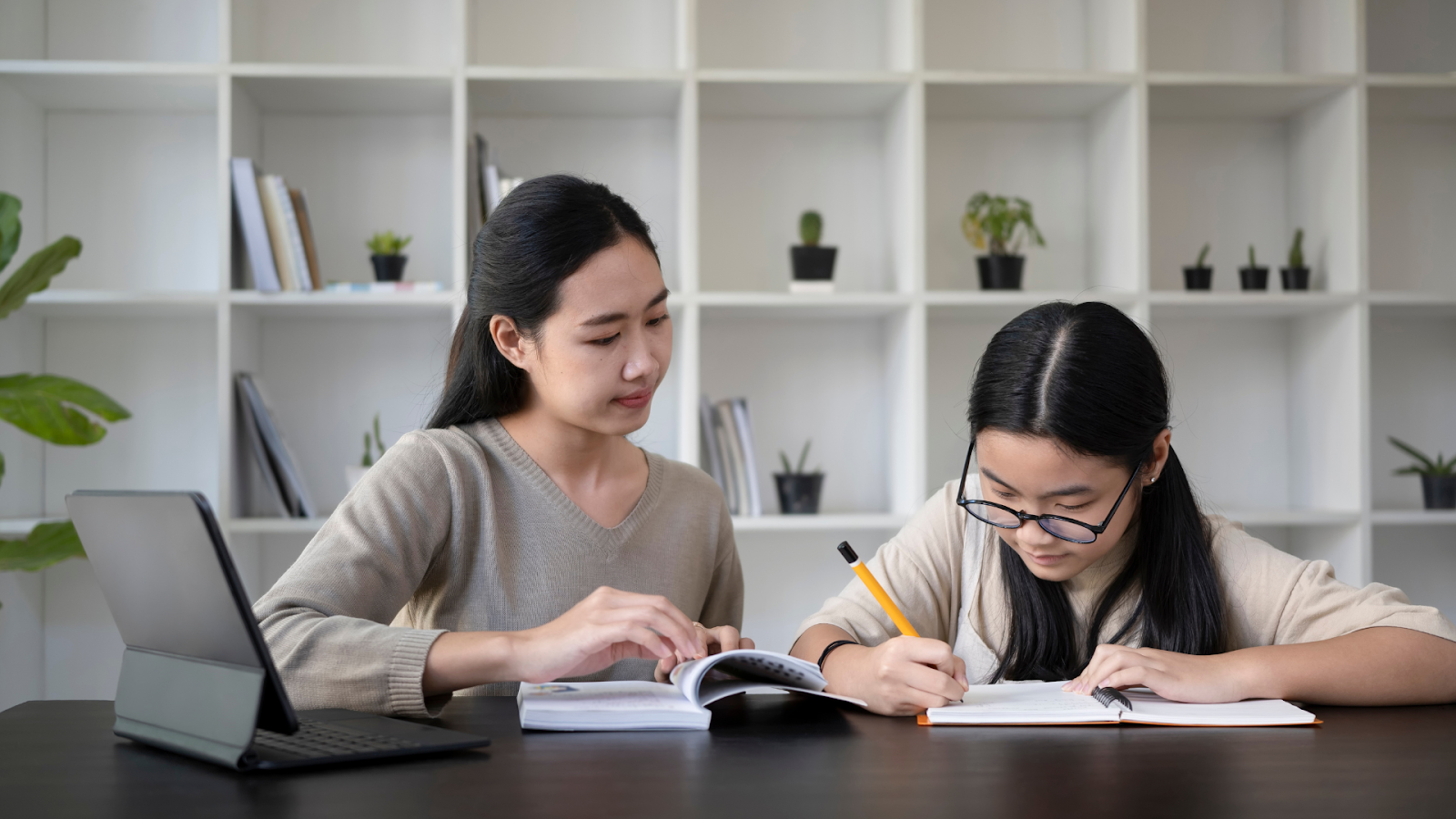 Two Asian women studying together at a table with books, notebooks, and a tablet in a modern room with white bookshelves and small potted plants in the background.