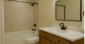 A modern 5×7 bathroom with a bathtub, shower, toilet, and vanity featuring a white countertop, wooden cabinet, black fixtures, and a large mirror against beige tiled walls.