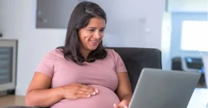 Pregnant woman smiling while working on laptop at home with hand on baby bump