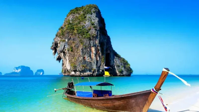 aditional Thai long-tail boat anchored in turquoise waters near limestone cliffs, showcasing scenic Thailand as featured in Flamingo Transworld tour packages.