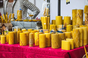 Display of golden yellow beeswax candles with decorative patterns on red tablecloth at outdoor artisan market, with vendor in striped sweater in background