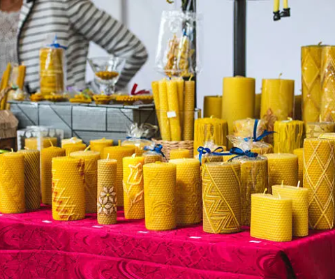 Display Of Golden Yellow Beeswax Candles With Decorative Patterns On Red Tablecloth At Outdoor Artisan Market, With Vendor In Striped Sweater In Background