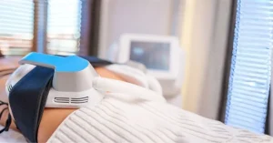 Close-up of an EMS Sculptor device treatment on a person's body, highlighting the non-invasive applicator secured with a strap, with a digital control screen visible in the soft-focus background.