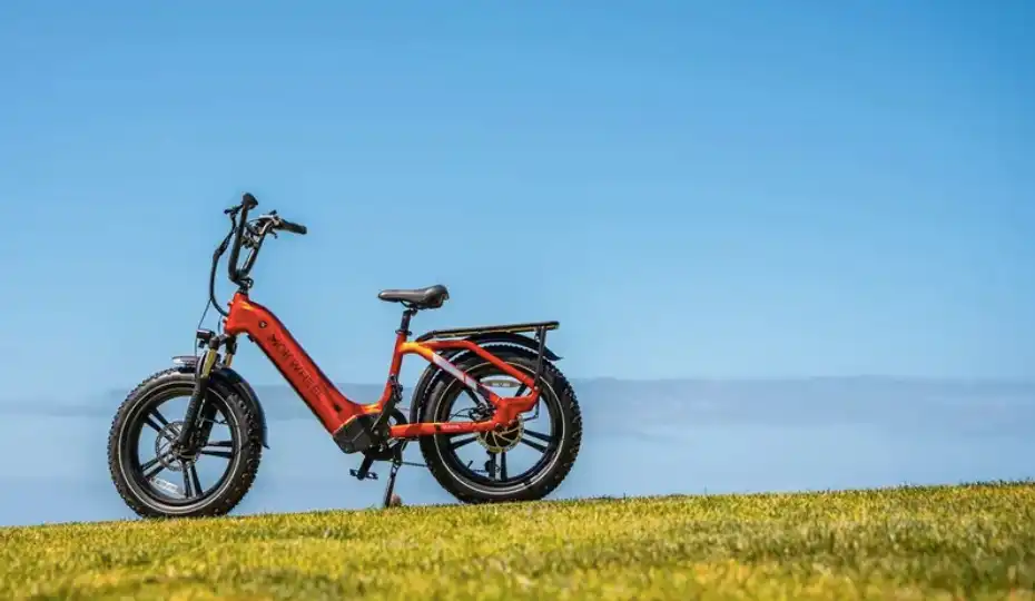 Bright red step-through electric bike with fat tires, rear cargo rack, and low frame parked on a grassy hill against a clear blue sky, showcasing its easy-access design and adventure-ready style.