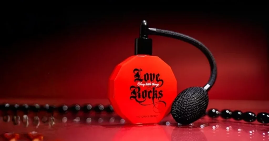 A striking red perfume bottle, octagonal, with a black atomizer bulb and hose, featuring "Love Rocks" in bold script, set against a deep red, reflective background with black beaded jewelry.
