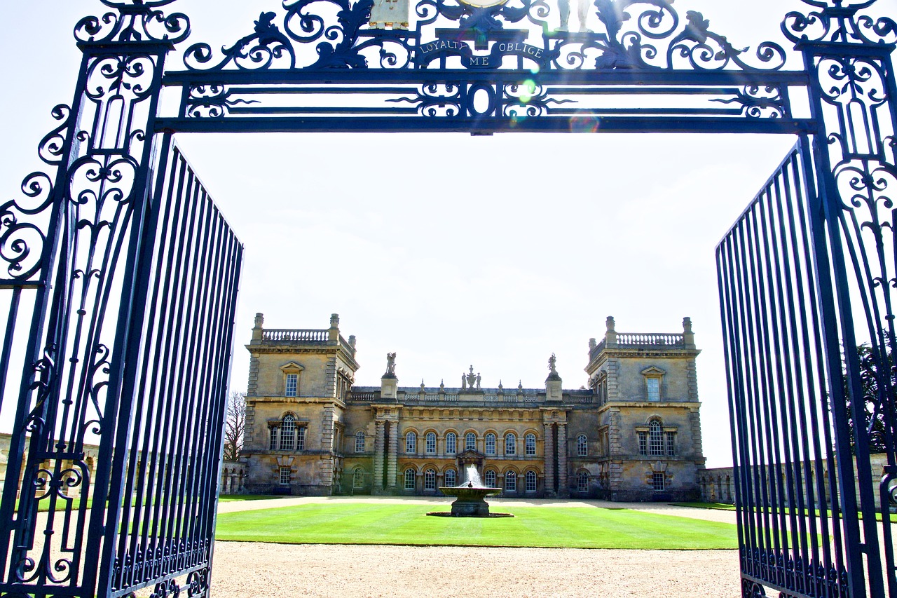 Ornate wrought iron automatic driveway gates with decorative scrollwork opening to historic estate mansion with manicured lawn
