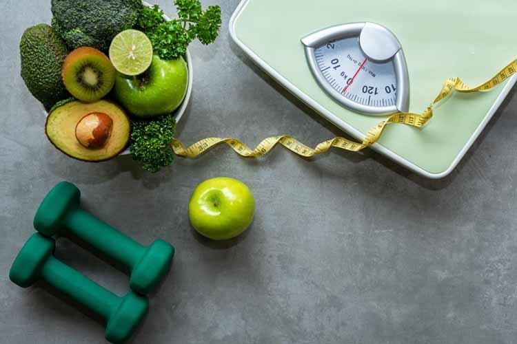 Healthy green foods including avocado, kiwi, lime, apple, broccoli, and parsley arranged with a bathroom scale, measuring tape, and green dumbbells, symbolizing sustainable weight loss through balanced nutrition and exercise.