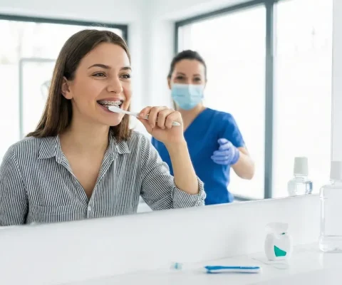 A Smiling Young Woman With Metal Braces Brushes Her Teeth In Front Of A Large Mirror, While A Masked Dental Professional In Blue Scrubs Observes Her In The Background Reflection.