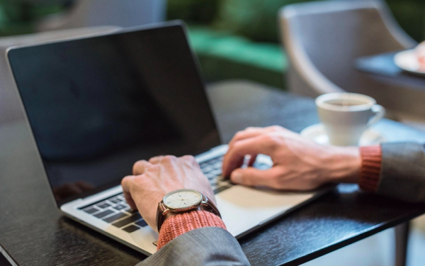 Person Typing On Laptop At Coffee Shop With Watch On Wrist And Coffee Cup Nearby