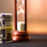A Roll Of Us Dollar Bills Secured With A Yellow Rubber Band Placed Beside A Wooden Hourglass On A Dark Surface