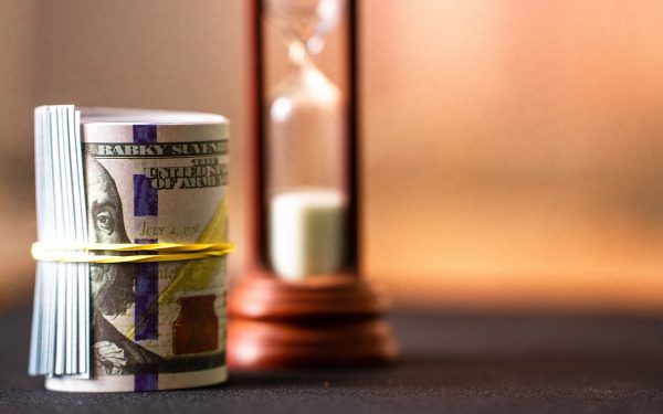 A Roll Of Us Dollar Bills Secured With A Yellow Rubber Band Placed Beside A Wooden Hourglass On A Dark Surface