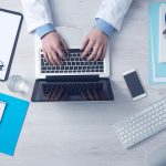 Overhead View Of A Doctor In A White Coat Typing On A Laptop At A Desk Surrounded By A Stethoscope, Smartphone, Keyboard, Clipboard, Notepad, Thermometer, And Medical Documents
