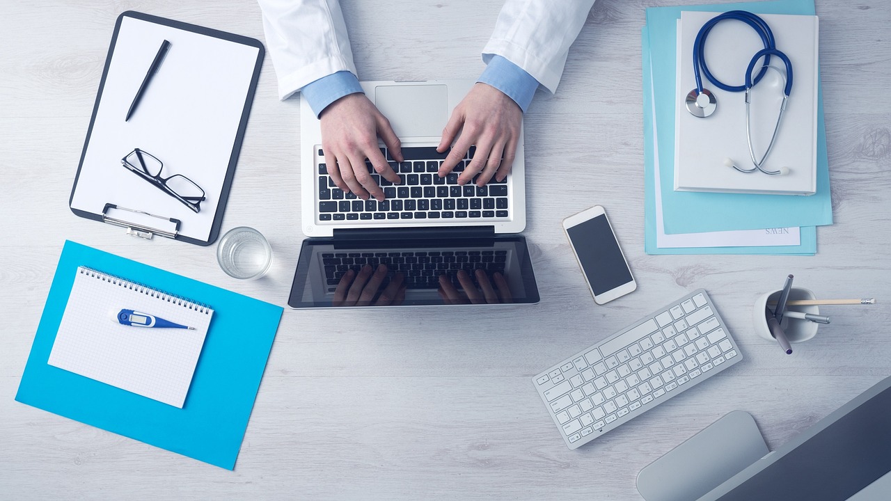 Overhead view of a doctor in a white coat typing on a laptop at a desk surrounded by a stethoscope, smartphone, keyboard, clipboard, notepad, thermometer, and medical documents
