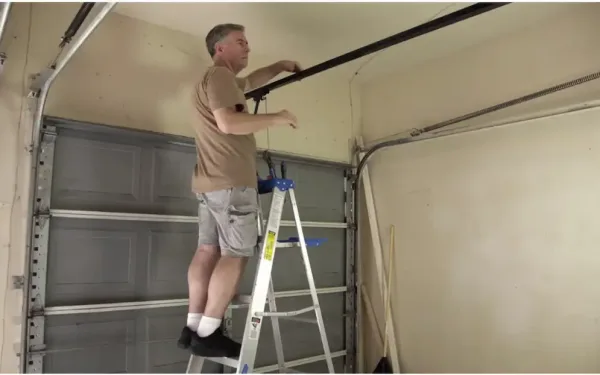 Technician Standing On A Ladder Inspecting The Overhead Garage Door Track And Hardware Inside A Garage